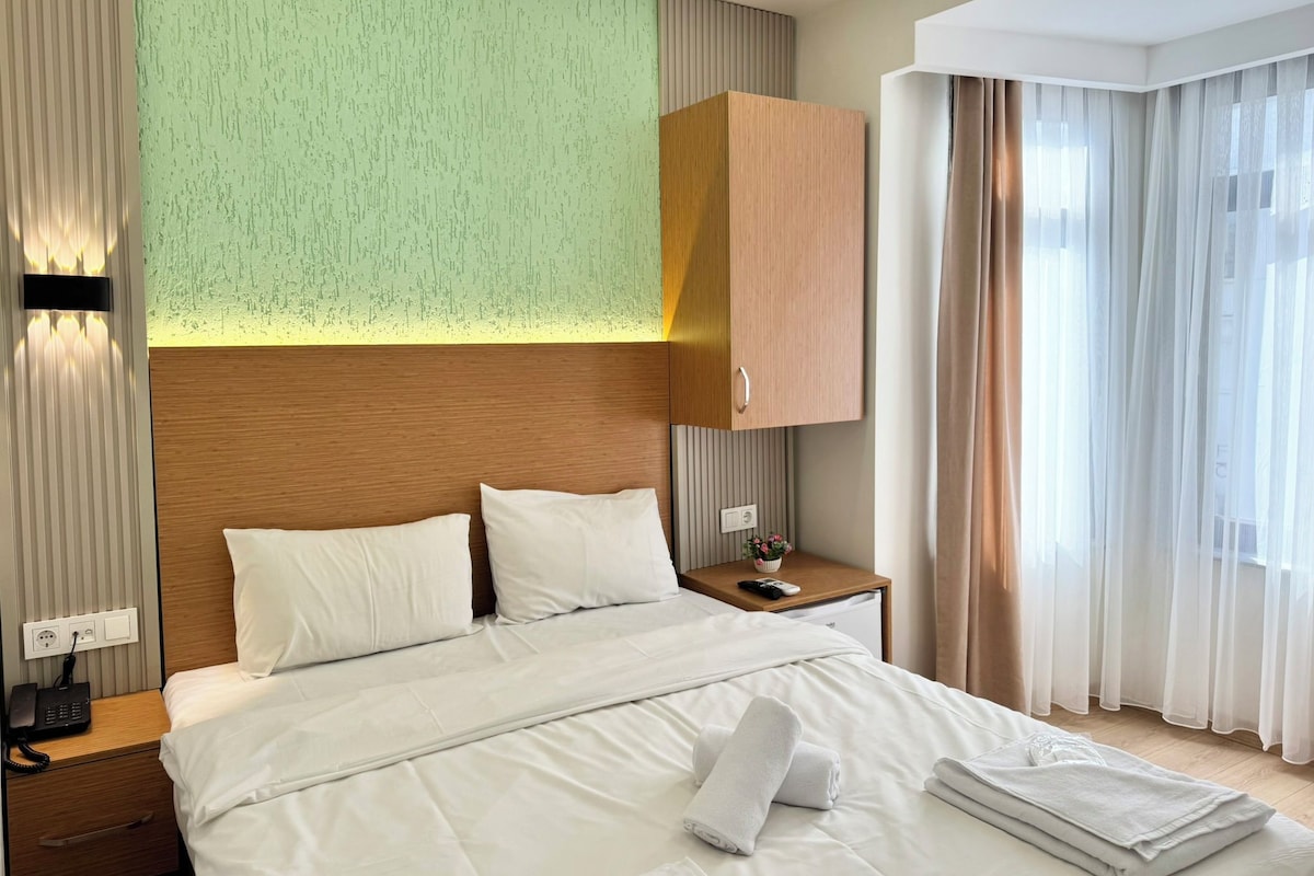 Quarto de hotel moderno com uma cama grande, cabeceira de madeira, mesa de cabeceira com telefone e vista para o exterior através de cortinas finas.