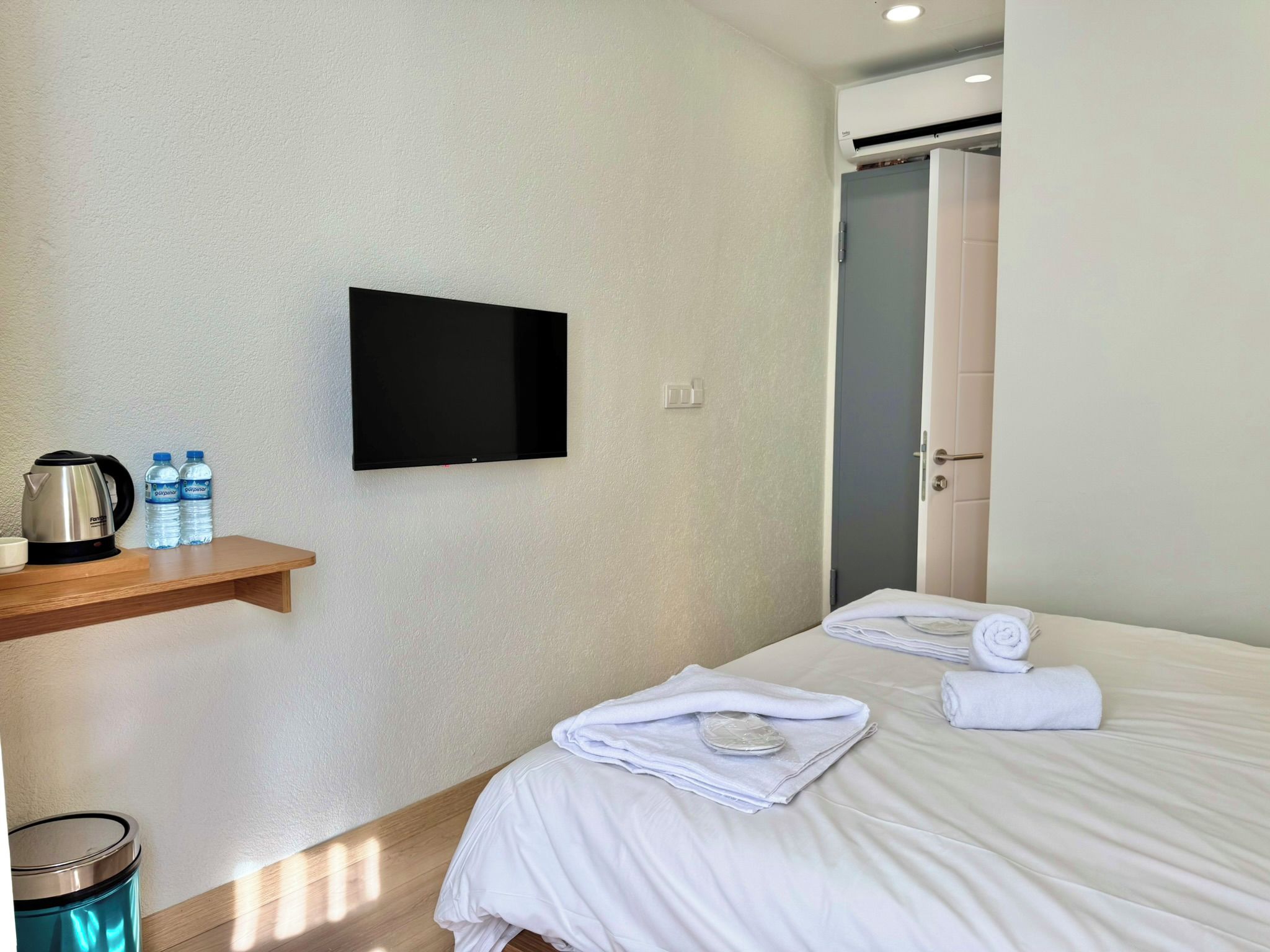 A hotel room with a bed, a television mounted on the wall, a shelf with a kettle and water bottles, and a trash bin.