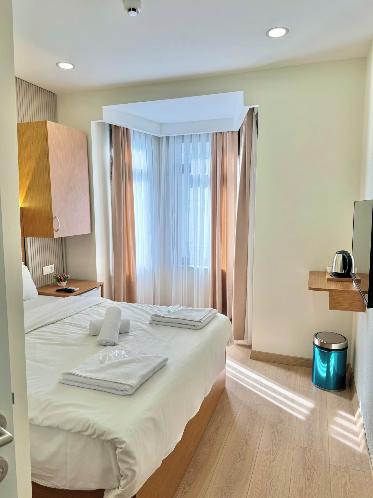 A hotel room with a bed, a nightstand, a TV, a kettle, and a trash bin.
