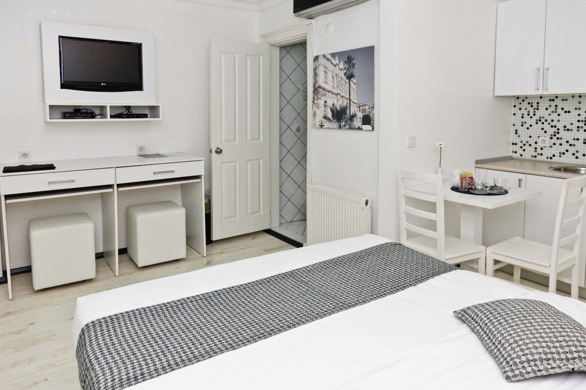 A modern hotel room with a bed, a desk, a television, and a small kitchenette.