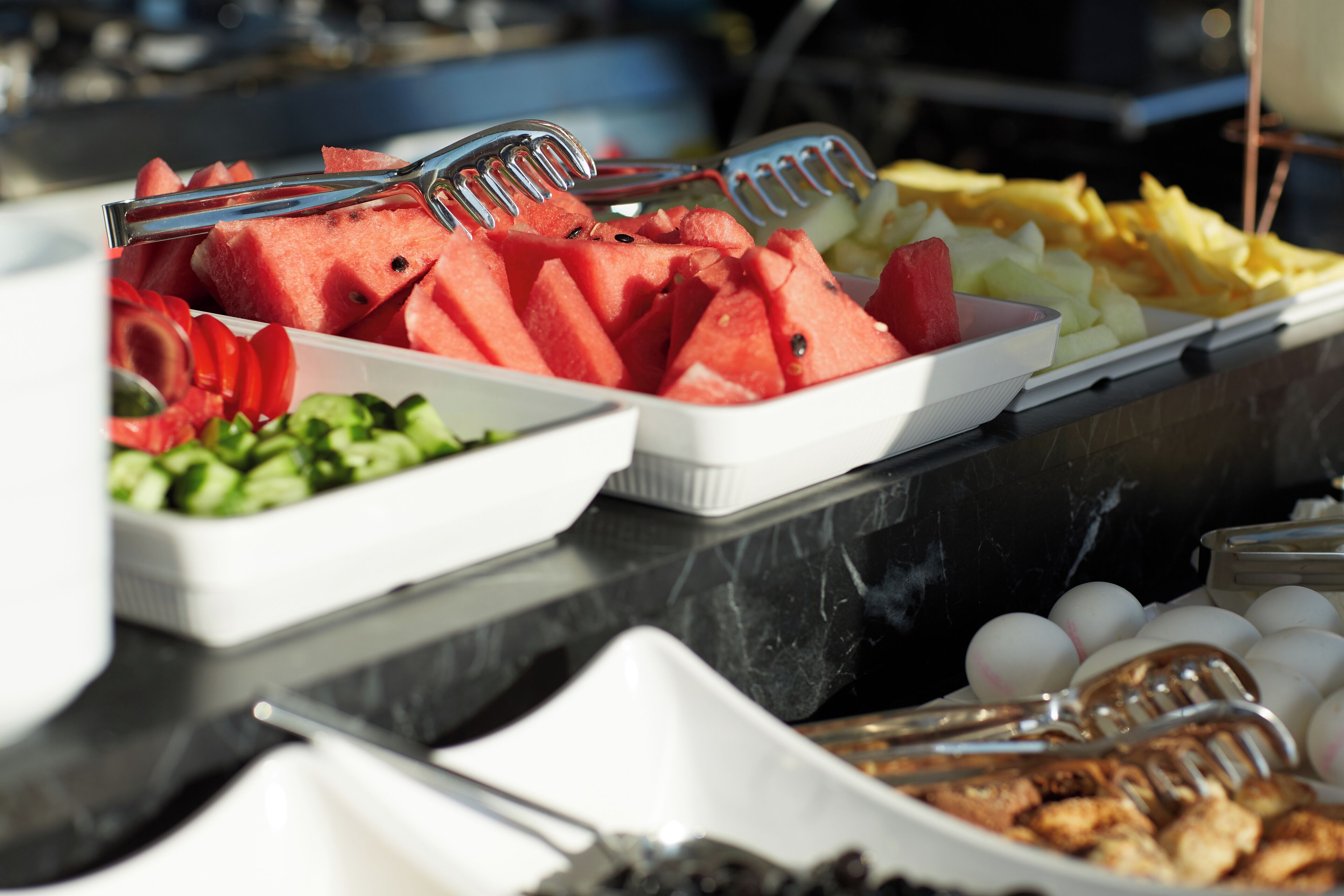 Un bufé con distintos tipos de comida, incluyendo sandía, ensalada, huevos y alimentos fritos.