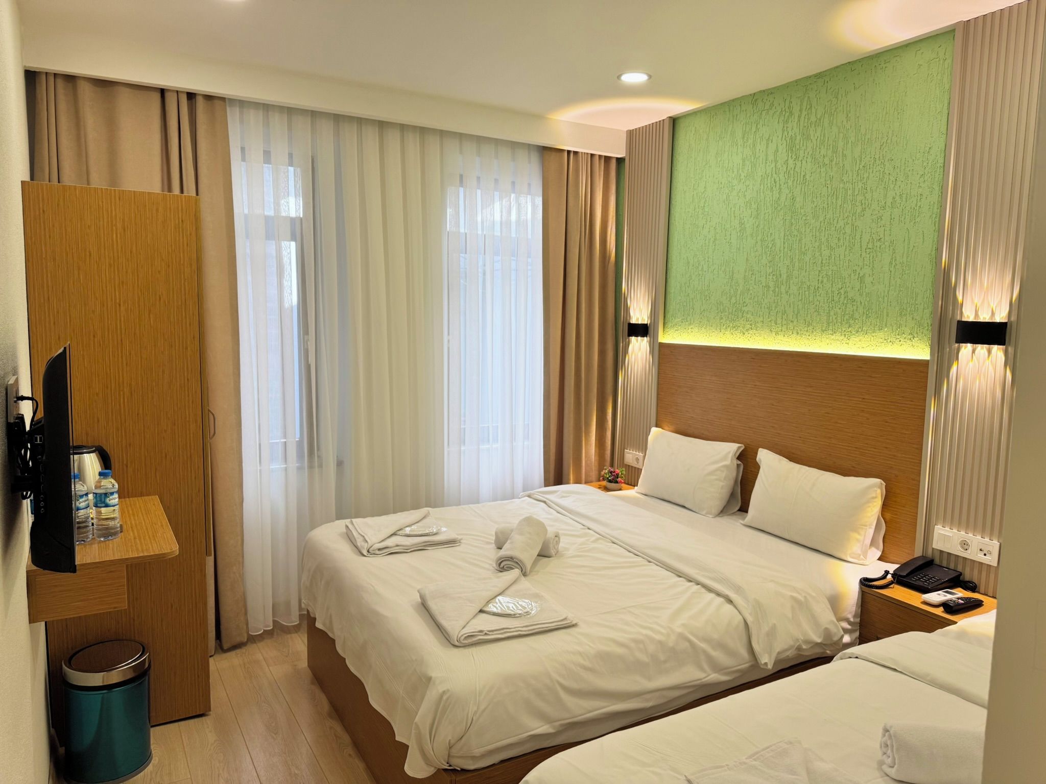 Ein Hotelzimmer mit zwei Betten, einem Holzschrank, einem Schreibtisch mit Telefon und Lampe sowie einer Wand in Grünton.