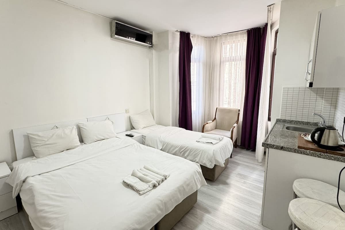 A hotel room with two beds, a chair, a small kitchen area, and a window with curtains.