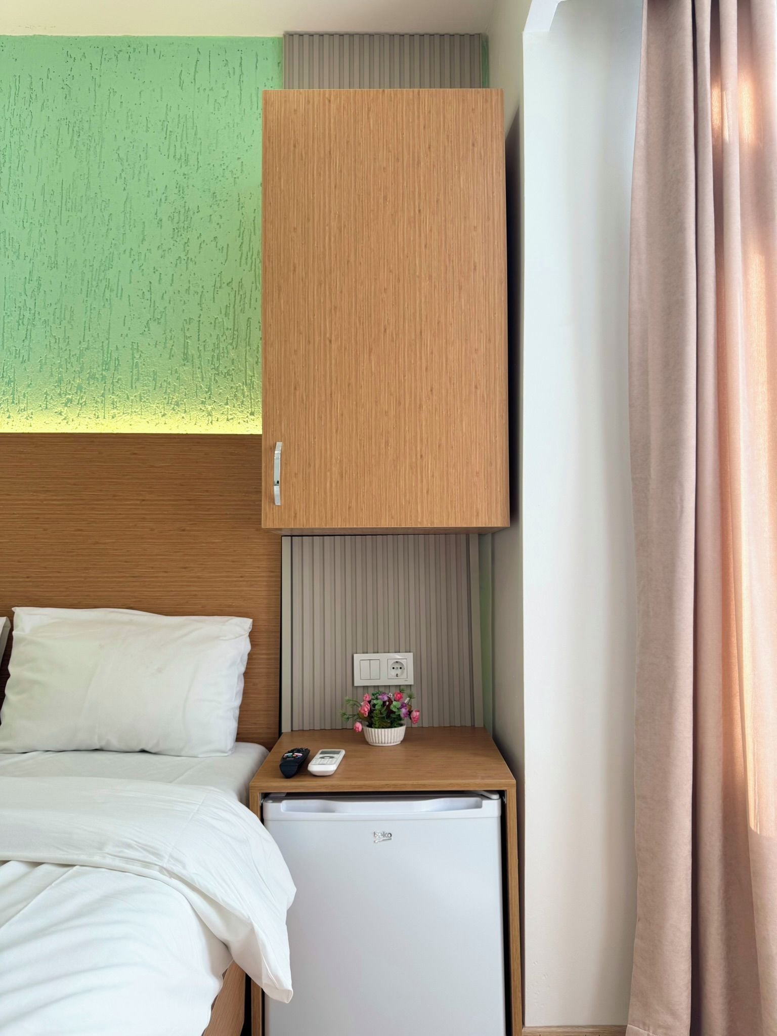 A hotel room with a bed, a small wooden cabinet, a mini fridge, a power outlet, and a potted plant.