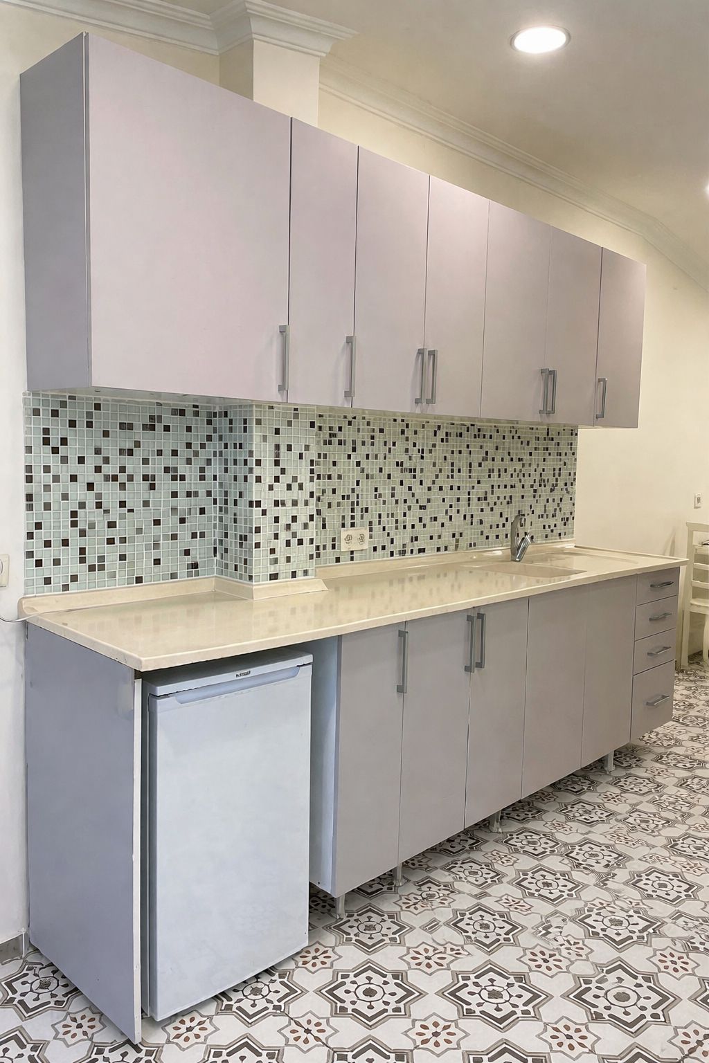A kitchen with a tiled backsplash, grey cabinets, a built-in dishwasher, and patterned floor tiles.
