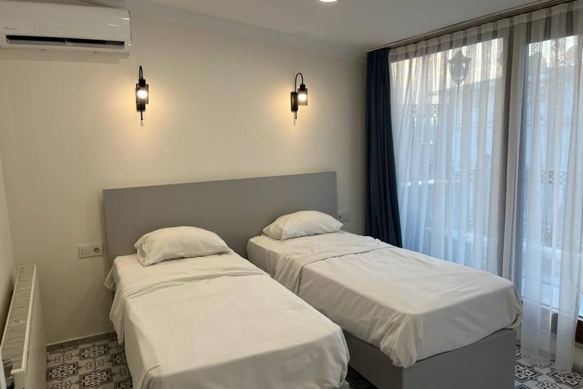 A hotel room with two single beds, a grey headboard, an air conditioner, and a window with sheer curtains.