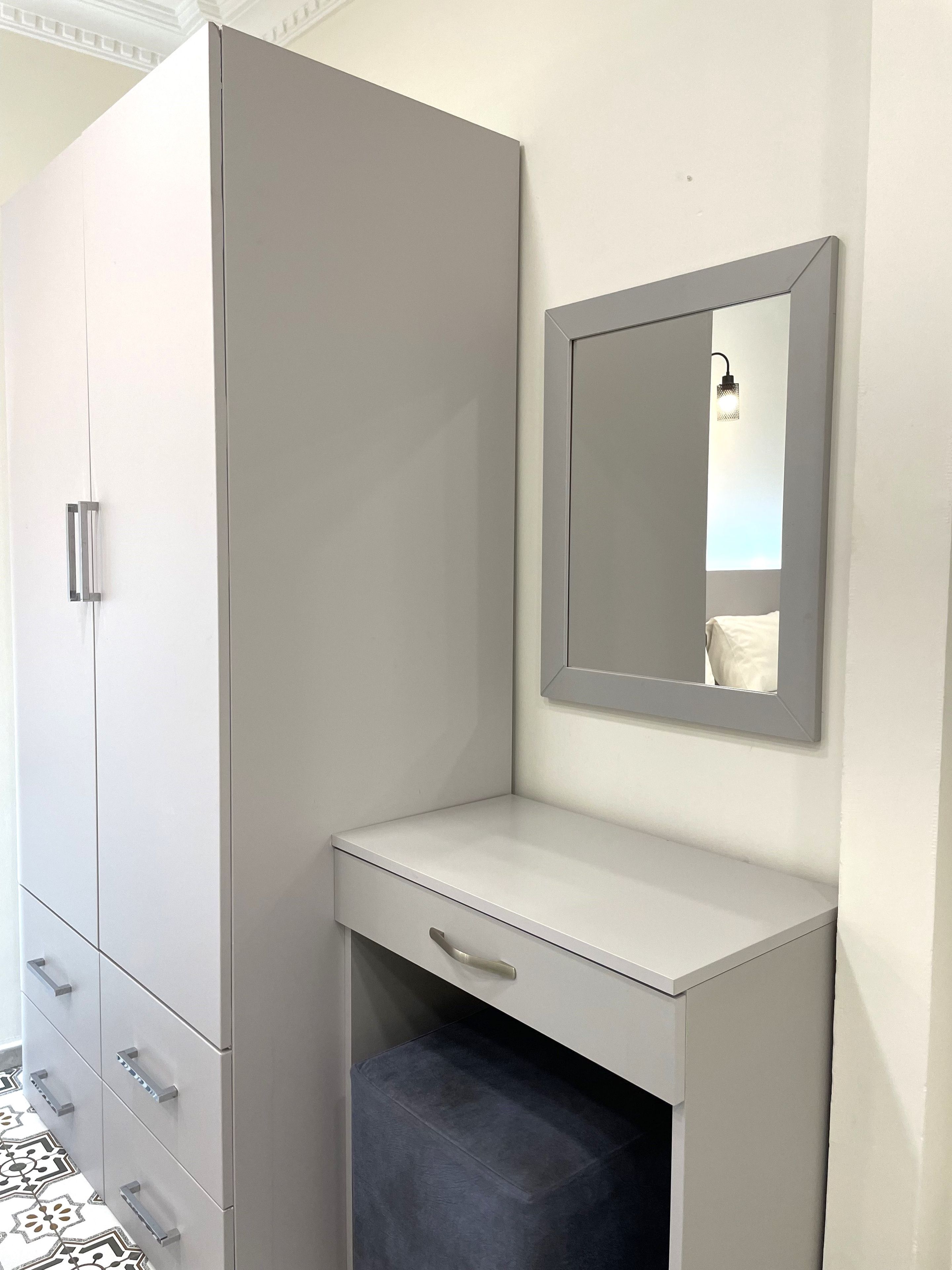A corner bedroom with a grey wardrobe, a bedside table, a mirror, and a patterned floor.