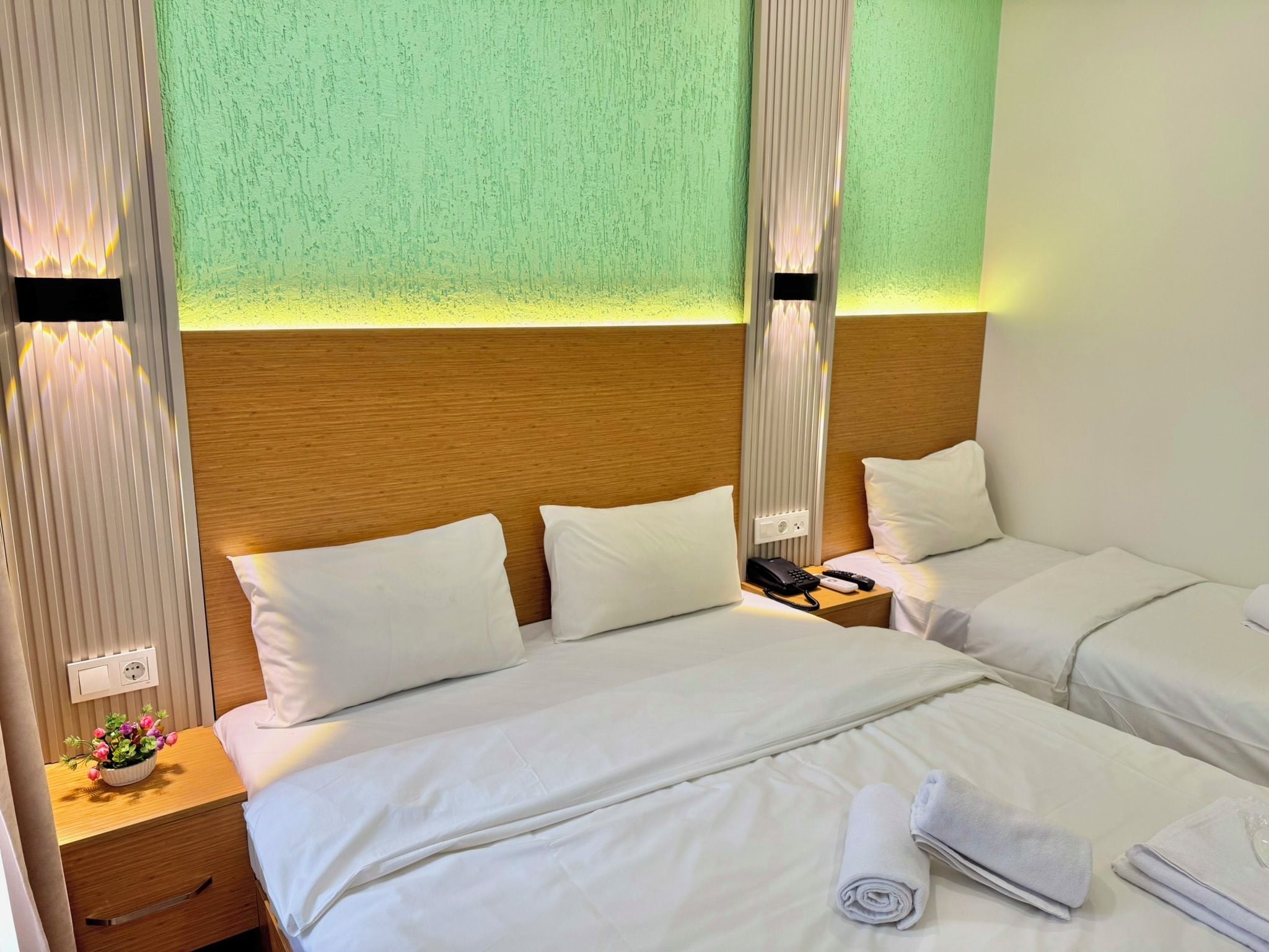 A hotel room with two beds, a wooden headboard, a nightstand with a phone and a lamp, and a wall-mounted outlet.