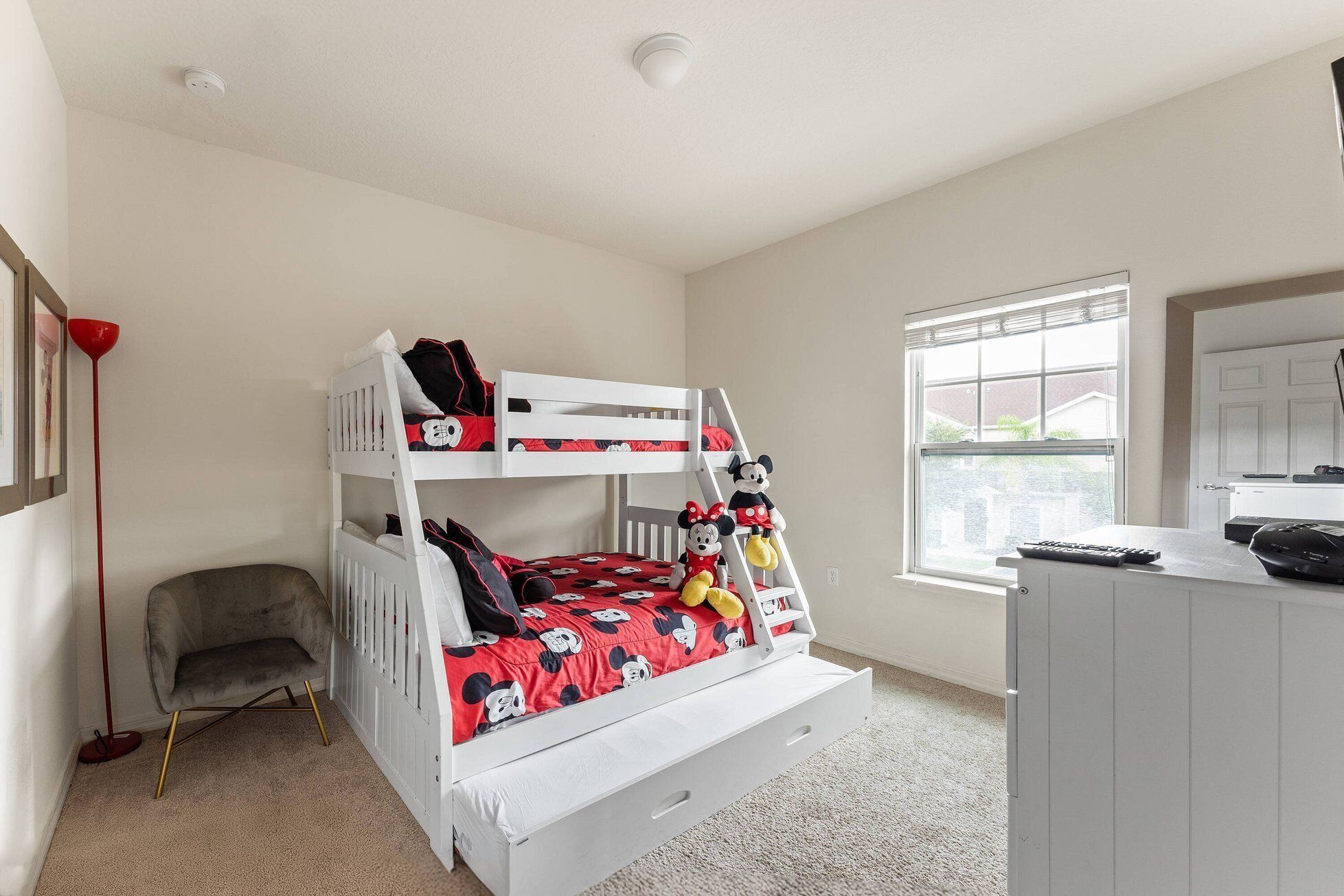 Un lit superposé avec une parure de lit aux motifs de Mickey Mouse, une chaise grise, une lampe de sol rouge et une fenêtre avec des stores.