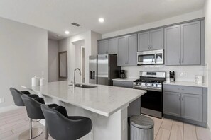 A modern kitchen with grey cabinets, a stainless steel refrigerator, a microwave, and a gas stove. There is a large island with a sink and bar stools. - 3BR Reunion Townhome, 10 min to Disney (Kissimmee)
