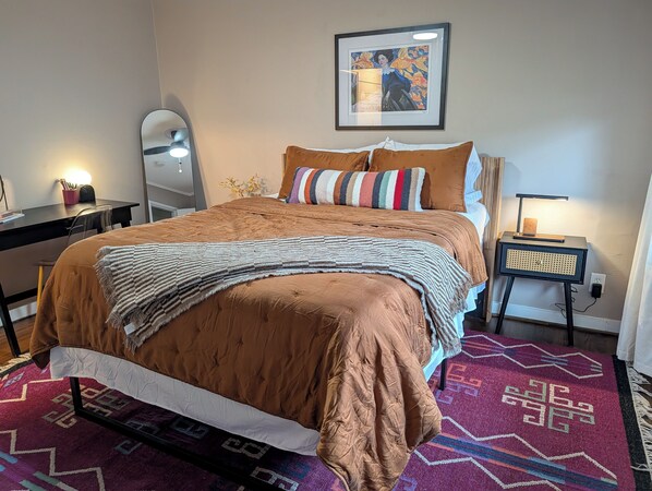 Een netjes ingerichte slaapkamer met een groot bed, een nachtkastje met een lamp, een kunstwerk in een lijst aan de muur, en een kamerkleed met een patroon.