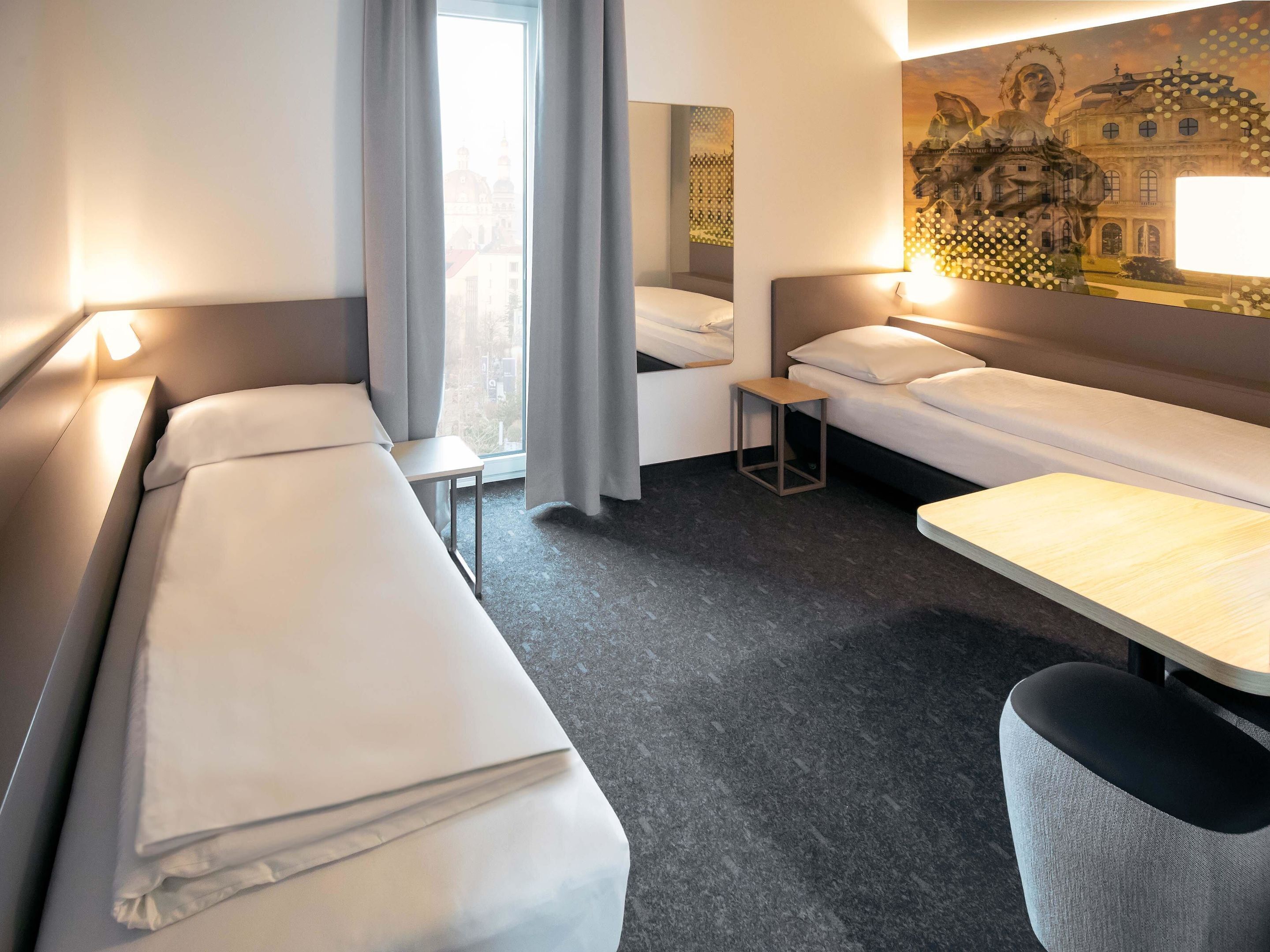 A hotel room with two single beds, a small table, a chair, and a large window with curtains.