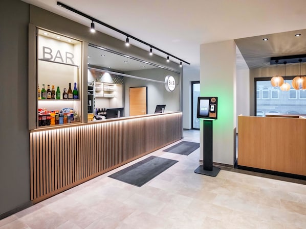 A modern hotel reception area with a bar, a reception desk, and a self-service kiosk.
