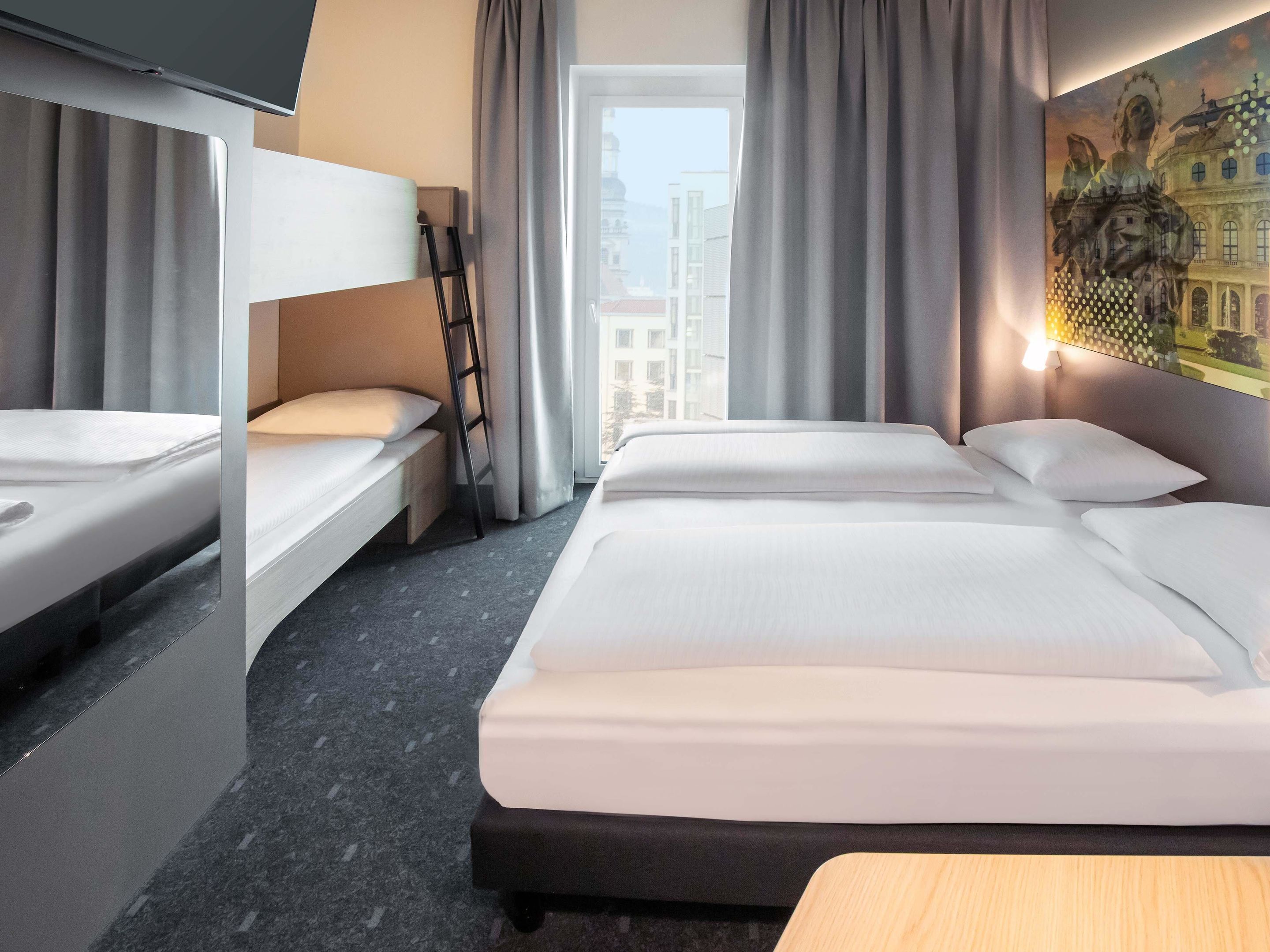 A hotel room with two beds, a bunk bed, a window with curtains, a painting on the wall, and a wooden bench.