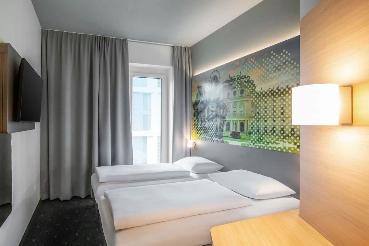 Ein Hotelzimmer mit zwei Betten, einem großen Fenster mit Vorhängen, einem Wandfernseher und einem Bild eines Gebäudes.