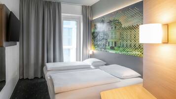 A hotel room with two beds, a large window with curtains, a wall-mounted TV, and a painting of a building.