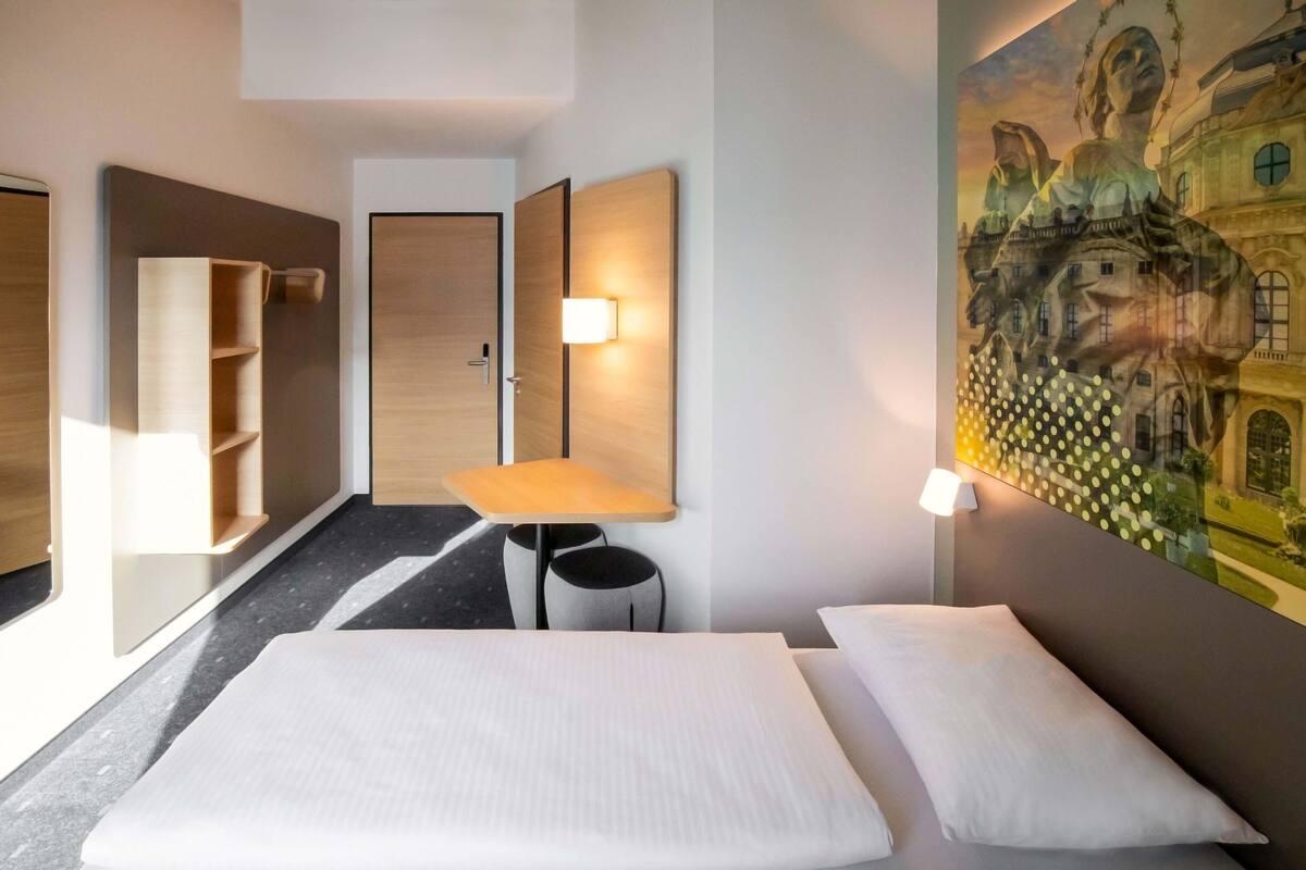 Ein Hotelzimmer mit Bett, Schreibtisch und Stuhl. An der Wand hängt ein Gemälde, im Hintergrund ist eine Tür zu sehen.