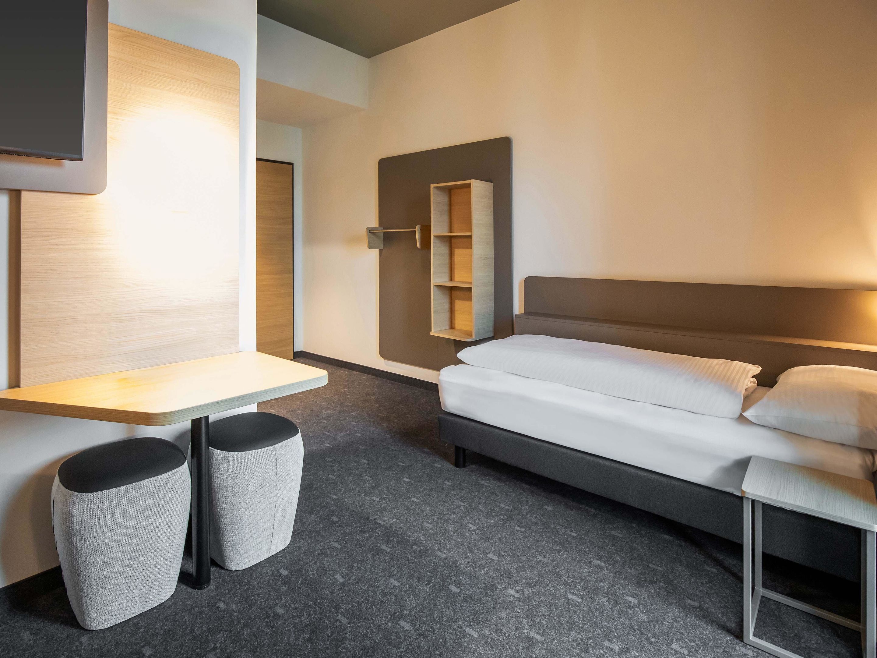 Una habitación de hotel moderna con una cama, un escritorio pequeño y una silla. Hay un espejo en la pared y un estante con algunos objetos.