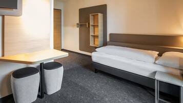 A modern hotel room with a bed, a small desk, and a chair. There is a mirror on the wall and a shelf with a few items.