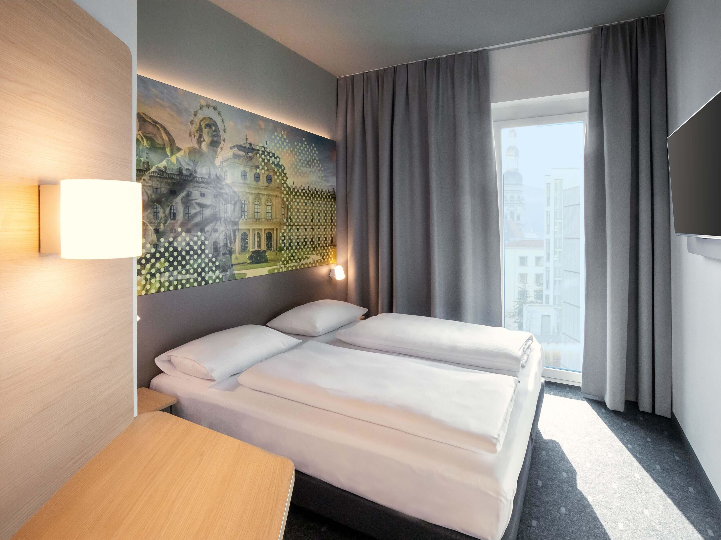 Habitación de hotel con cama, escritorio, lámpara, un cuadro en la pared y ventana con cortinas.