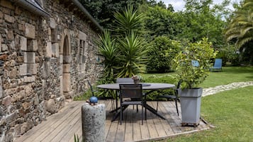 Un mur de pierre, une terrasse en bois, une table ronde avec des chaises et des plantes en pot.