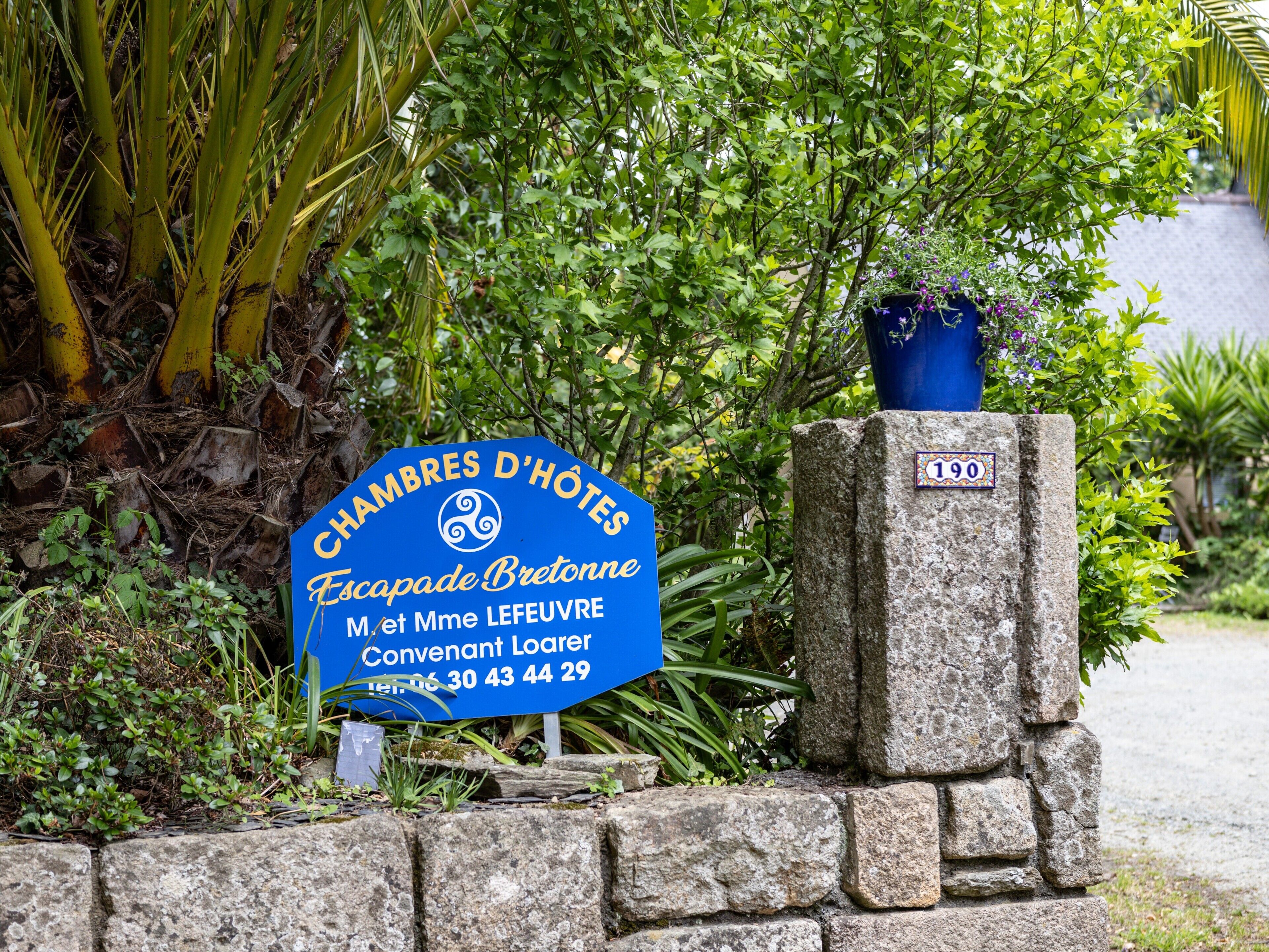 Une enseigne bleue indiquant « CHAMBRES D’HÔTES Escapade Bretagne », avec un pilier en pierre et une plante en pot.