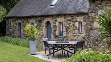 Een stenen gebouw met een betegeld dak, een terras met een tafel en stoelen, en potplanten.