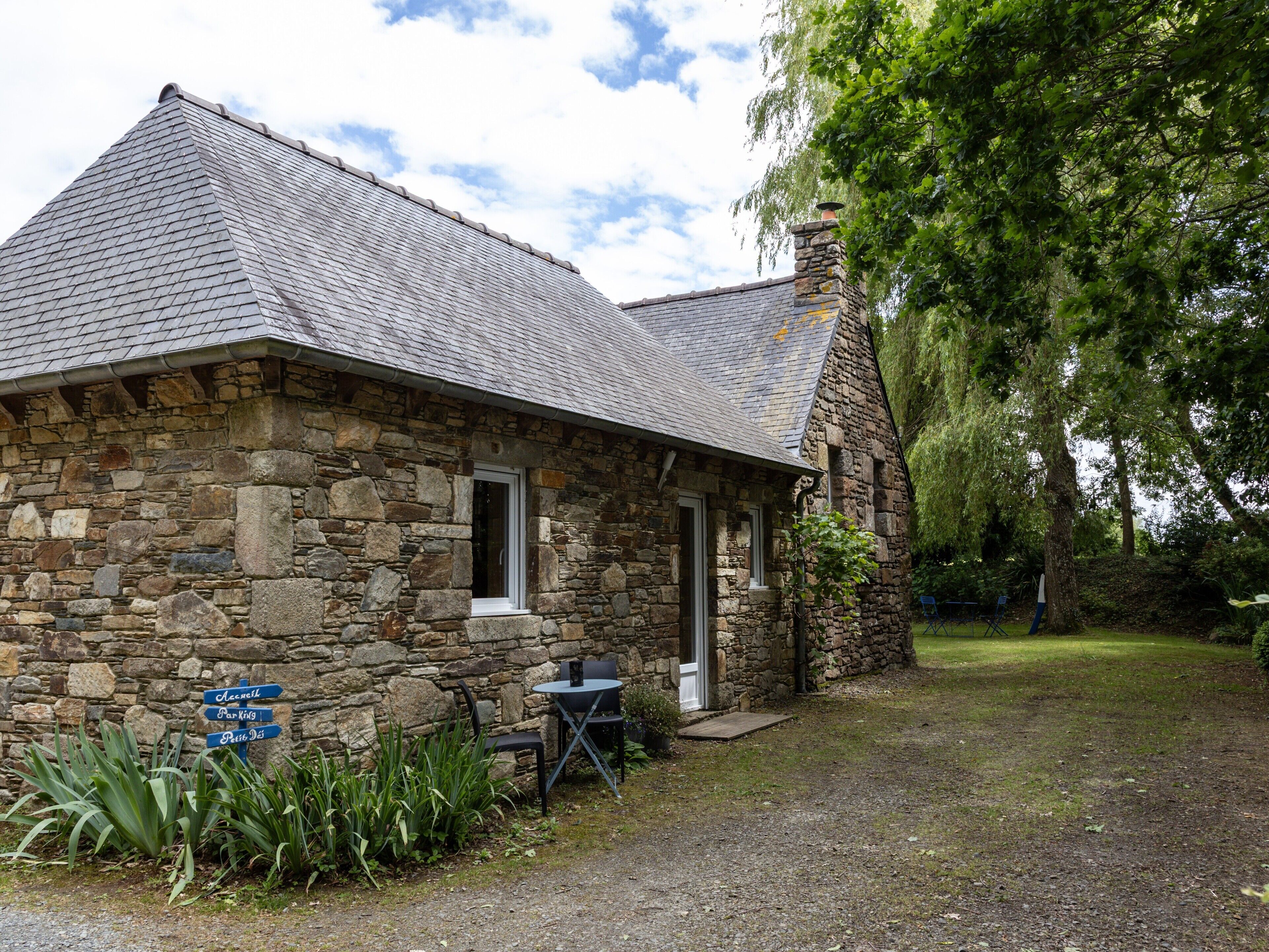 Un bâtiment en pierre avec un toit en pente, une porte et une petite table avec des chaises à l’extérieur.