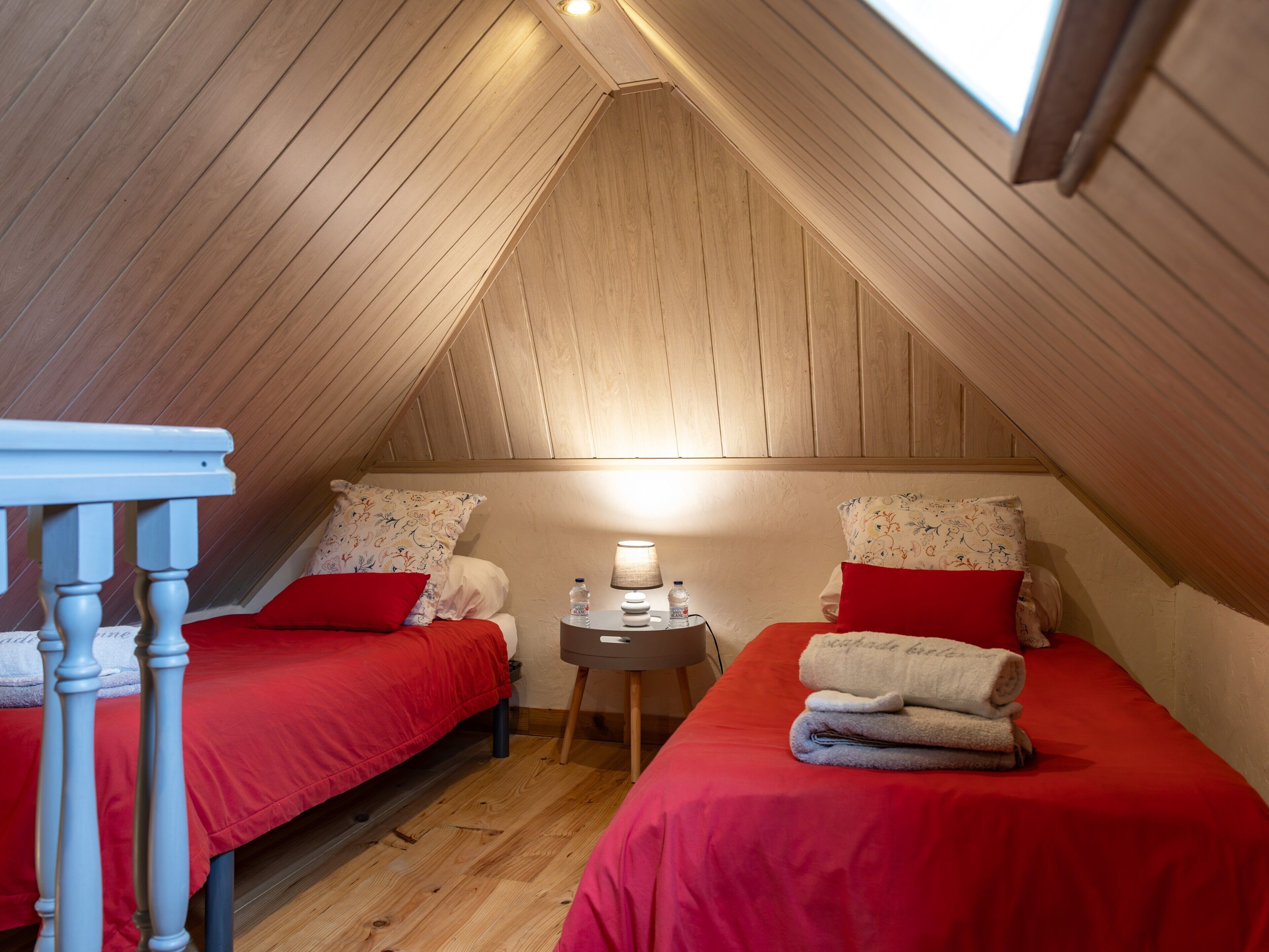 A small attic bedroom with two beds, a nightstand, a lamp, and a wooden ceiling.