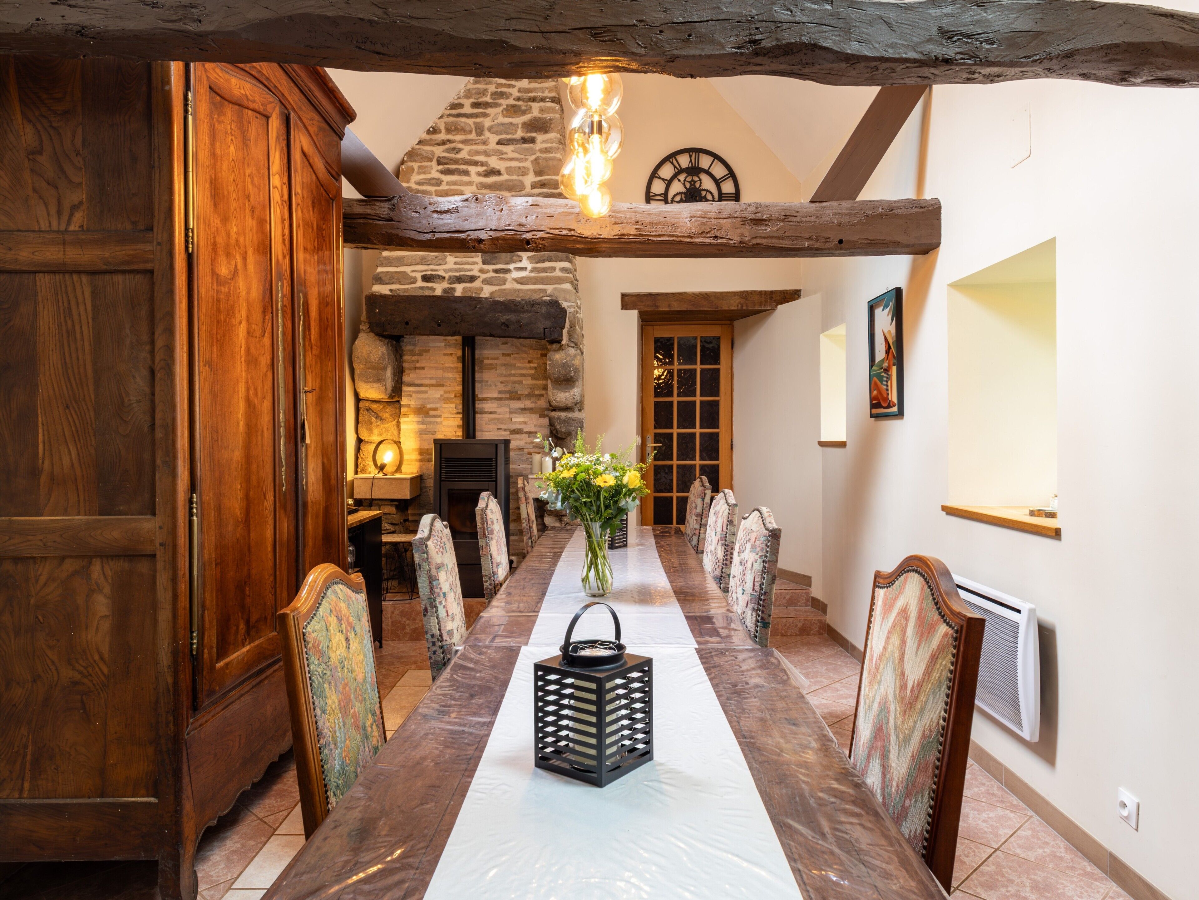 A dining area with a long table, chairs, a vase with flowers, a clock, and a fireplace.