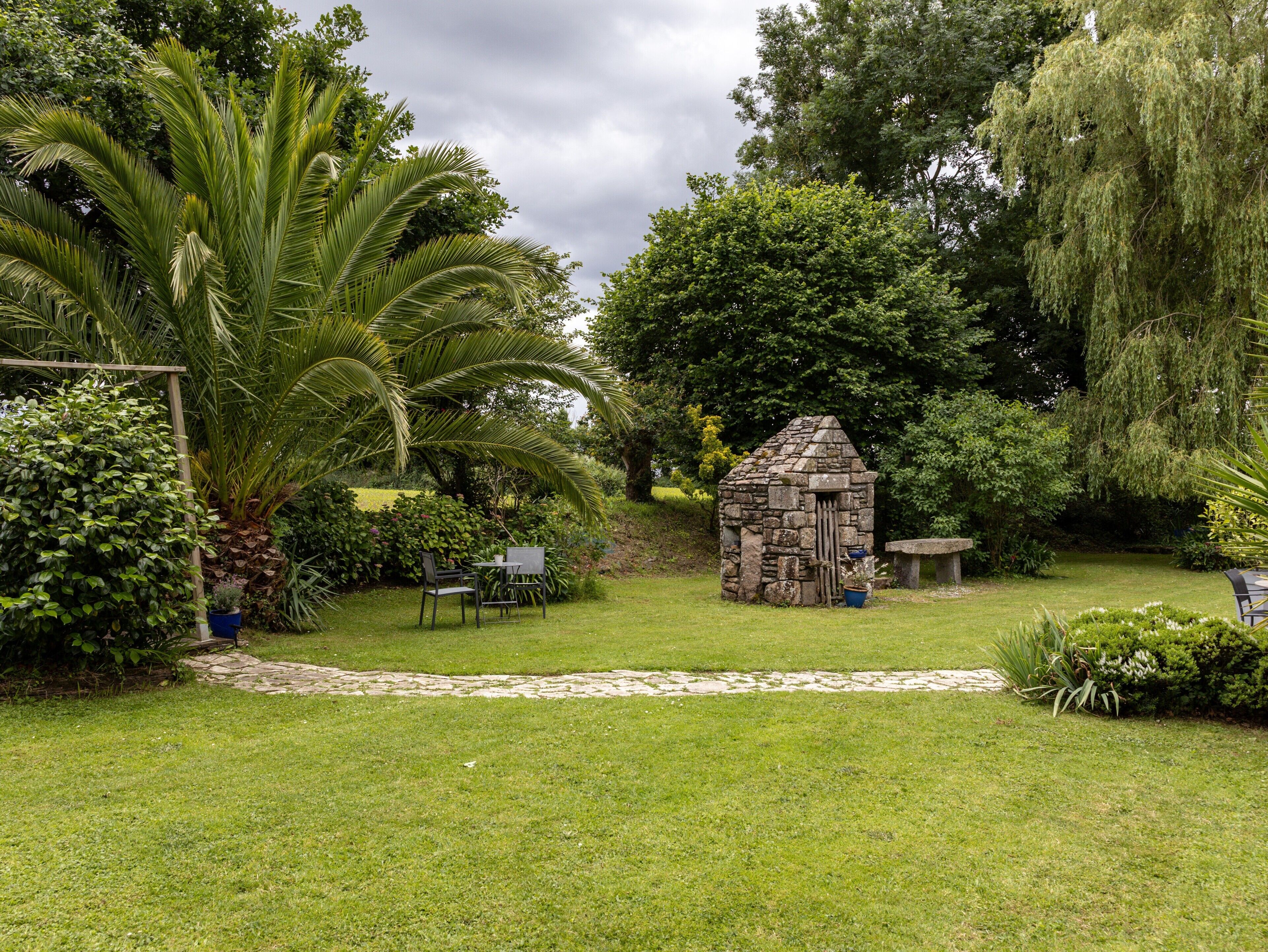 Une construction en pierre, située dans un jardin luxuriant, avec un sentier menant à celle-ci, et entourée de diverses plantes et arbres.
