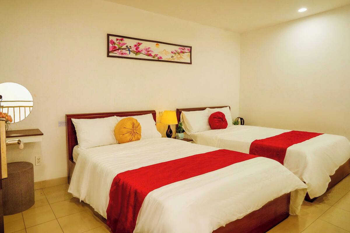 A hotel room with two beds, each with white bedding and red accents, a nightstand with a lamp, a wall-mounted painting, and a small trash bin.
