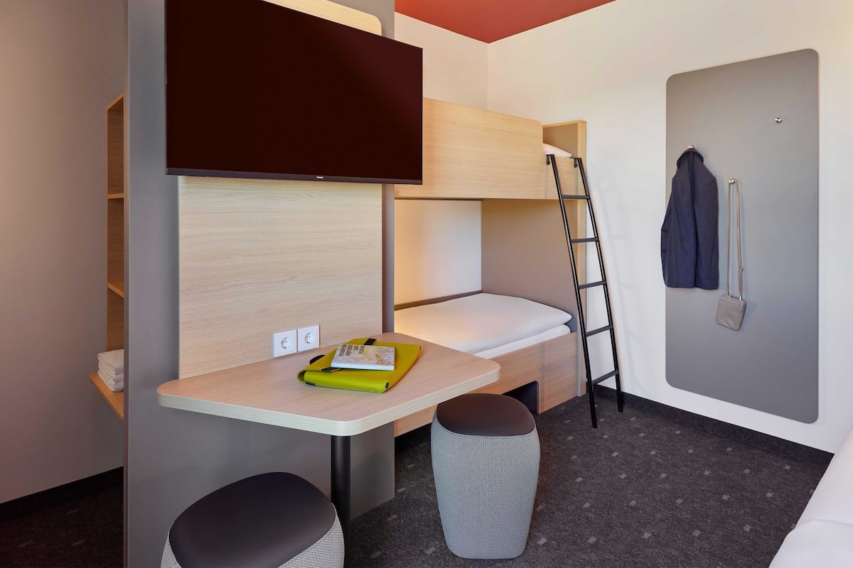 A compact hotel room with a bunk bed, a desk with a chair, a small stool, a wall-mounted TV, and a coat rack.
