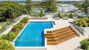 Una piscina rectangular con una cubierta de madera y escalones de acceso, rodeada de abundante vegetación y con vistas a una zona costera con edificios y un muelle.
