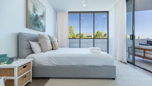 Un dormitorio moderno con una cama grande, una mesita de noche, un mueble de almacenamiento de mimbre y vistas a zonas verdes a través de un amplio ventanal.