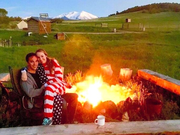 Un couple est assis près d’un feu de camp, avec un paysage montagneux en arrière-plan.
