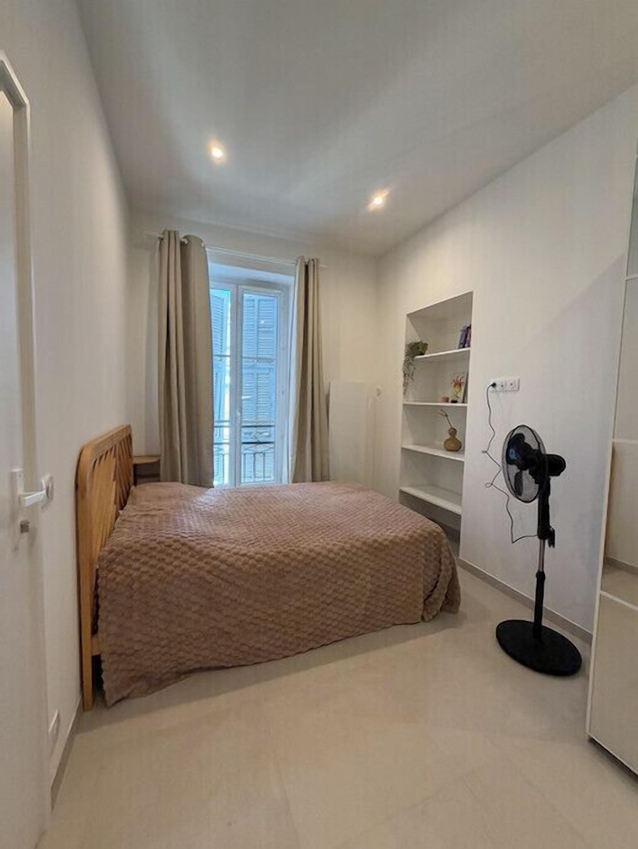 A bedroom with a bed, a standing fan, a window with curtains, and a white wall with shelves.