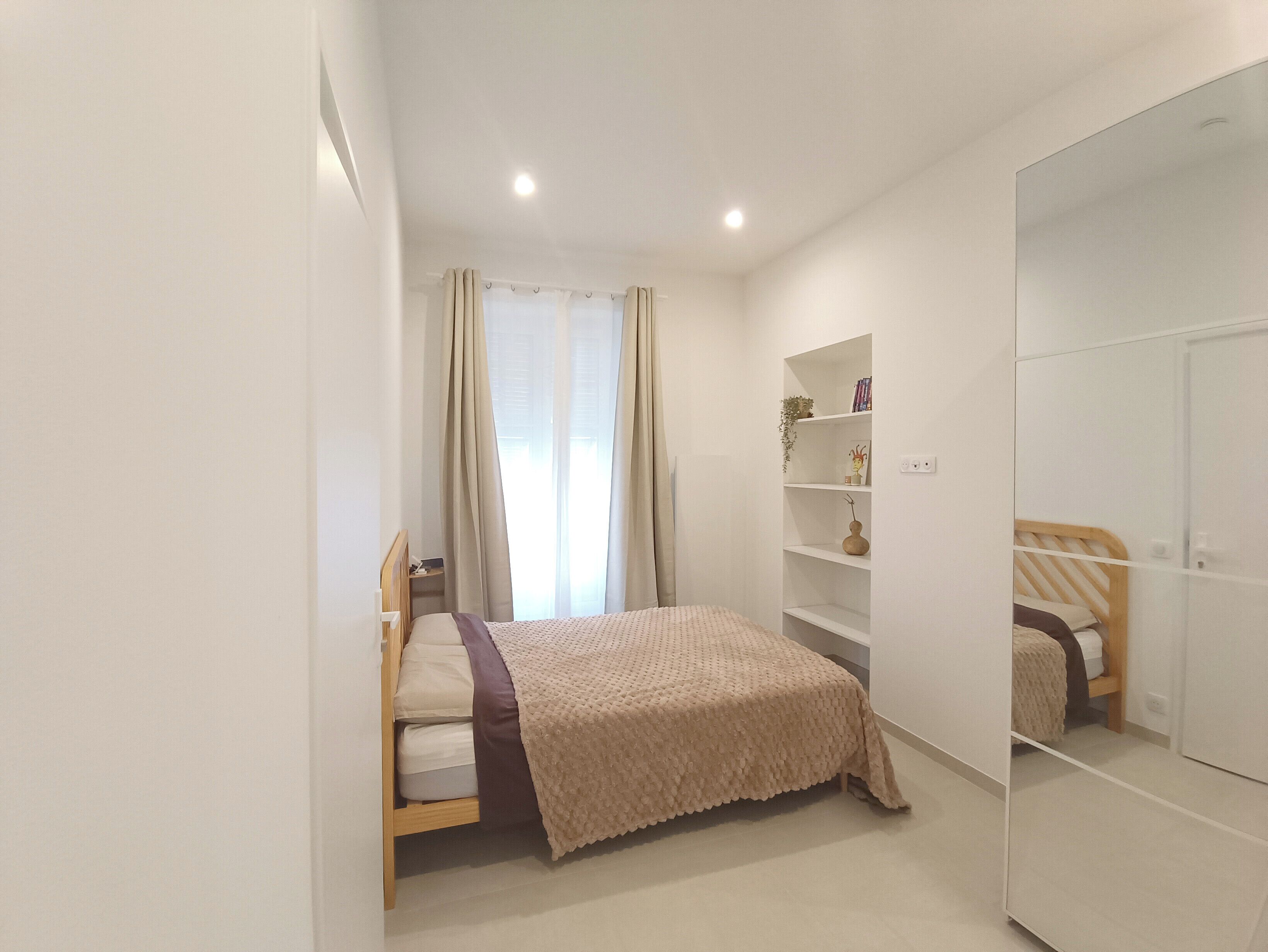 Une chambre à coucher avec un lit, une armoire, une étagère avec des livres et une plante, et une fenêtre avec des rideaux.