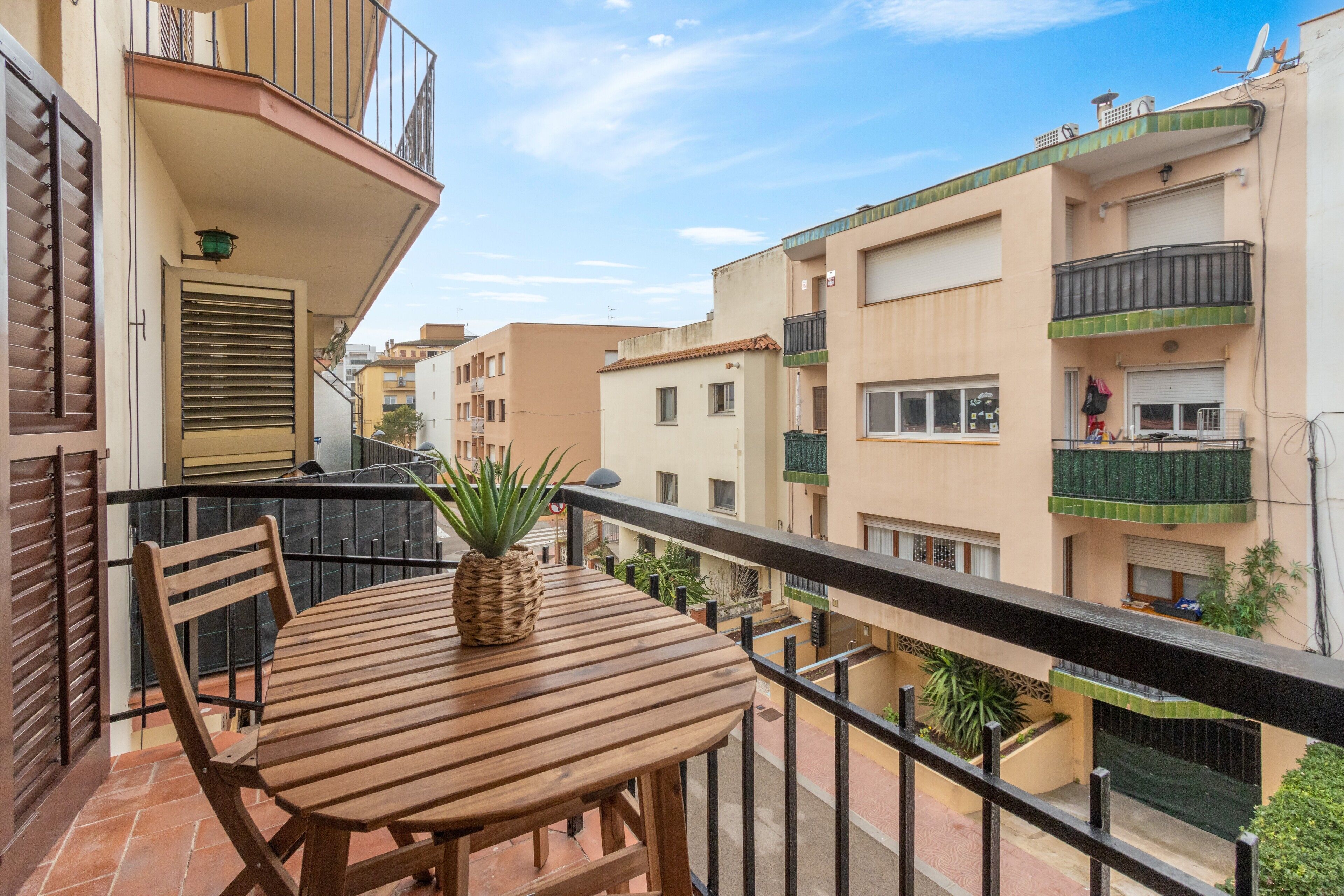 Un balcón con una mesa y sillas de madera, una planta en maceta y vistas a edificios cercanos.
