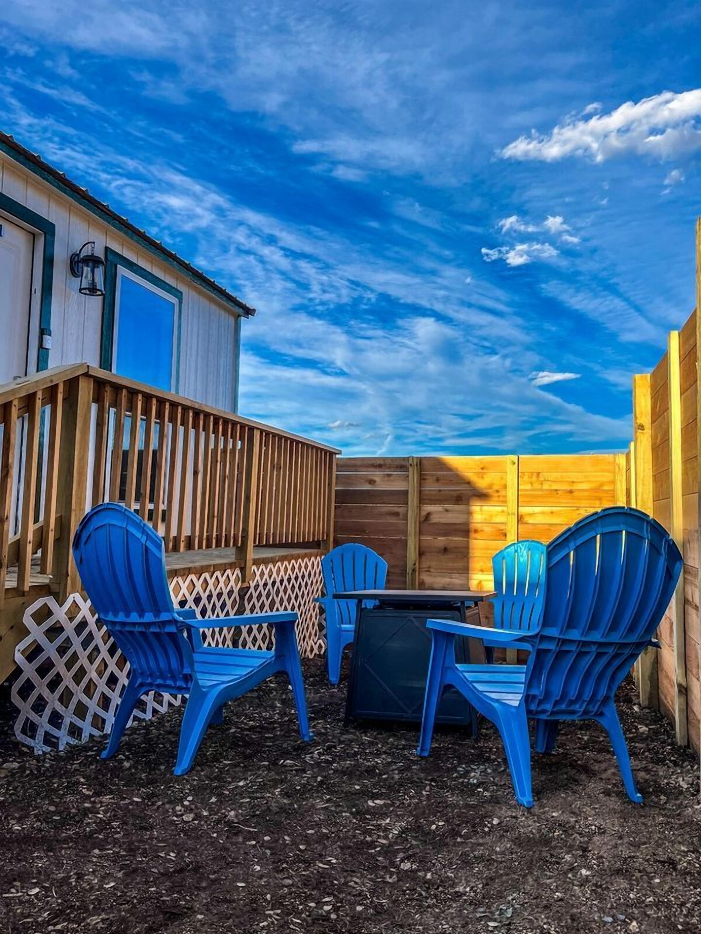 A patio with a table and four blue chairs, a wooden fence, and a building with a blue door.