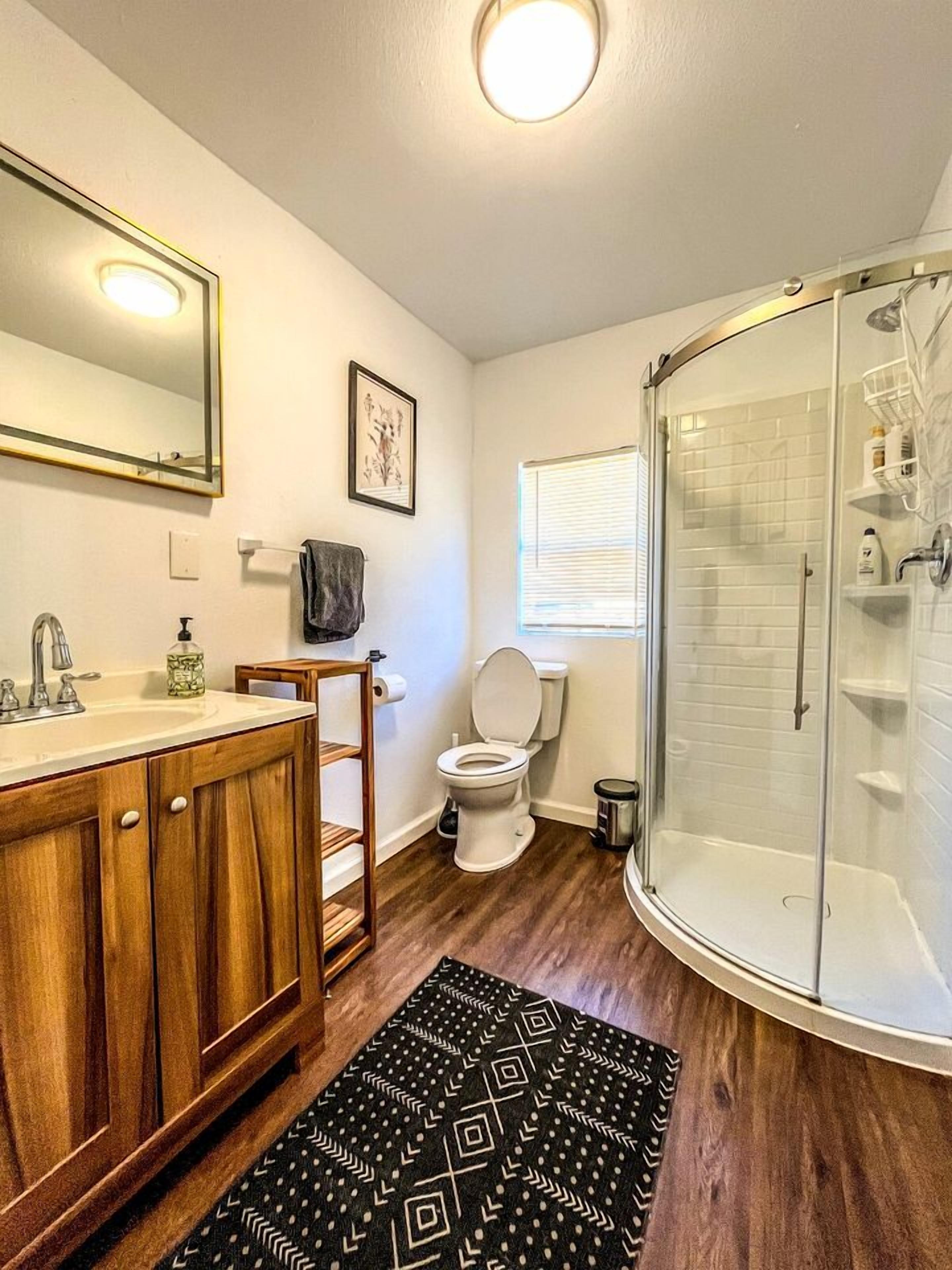 A bathroom with a wooden vanity, a toilet, a shower enclosure, and a patterned rug.