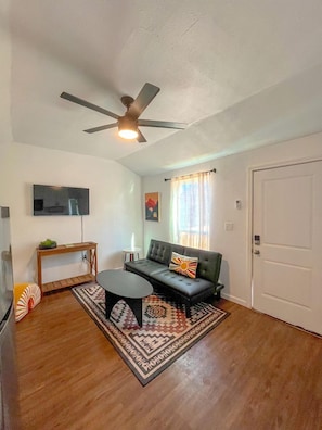 A living room with a ceiling fan, a sofa, a coffee table, a TV, and a door. - Cute Casita with Stock Tank Hot Tub and Fenced Yard in Lakehills, Texas (West Lake Hills)