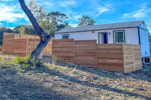 A small, single-story building with a metal roof, surrounded by wooden fences. - Cute Casita with Stock Tank Hot Tub and Fenced Yard in Lakehills, Texas (West Lake Hills)