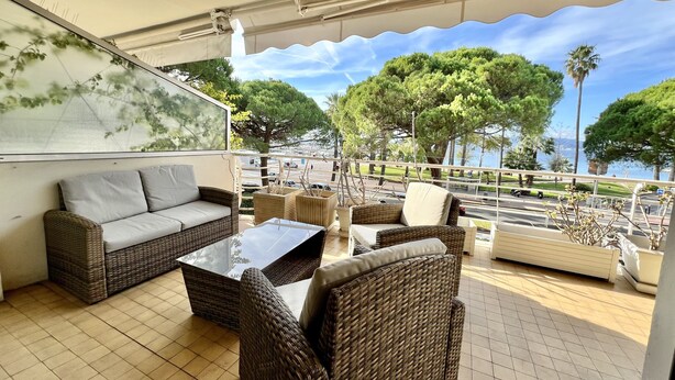 Un espace extérieur aménagé avec des meubles en osier, une table à plateau de verre et une vue sur la mer et les arbres.