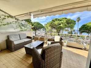 A covered outdoor seating area with wicker furniture, a glass-top table, and a view of the sea and trees. - Apt in Pointe Croisette, beach & large terrace (Cannes)