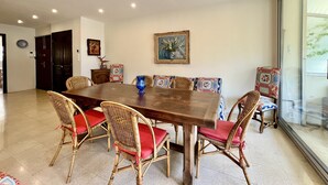 Un coin repas avec une grande table en bois entourée de chaises en osier dotées de coussins rouges. Un tableau orne le mur, et une fenêtre offre une vue sur la verdure.