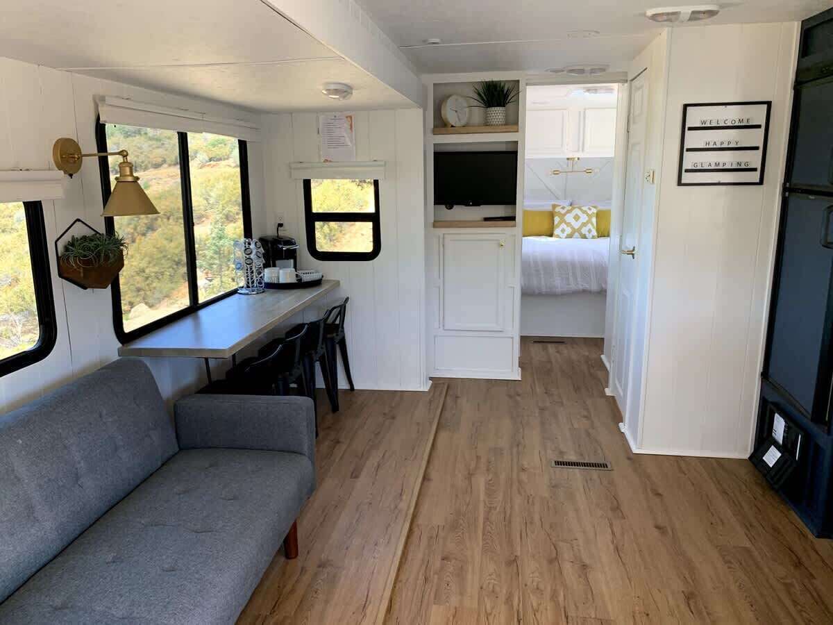 A compact RV interior with a gray sofa, a dining area, a kitchen with a TV, and a sleeping area with a bed.