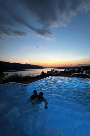 A swimming pool with two people in it, overlooking a body of water and mountains during sunset. - PENSIUNEA BERZASCA (Berzasca)