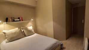 A hotel room with a bed, bedside lamps, a shelf with books, and a door leading to another room. - Apartment sea view in Saint-Raphaël w/pool (Saint-Raphaël)