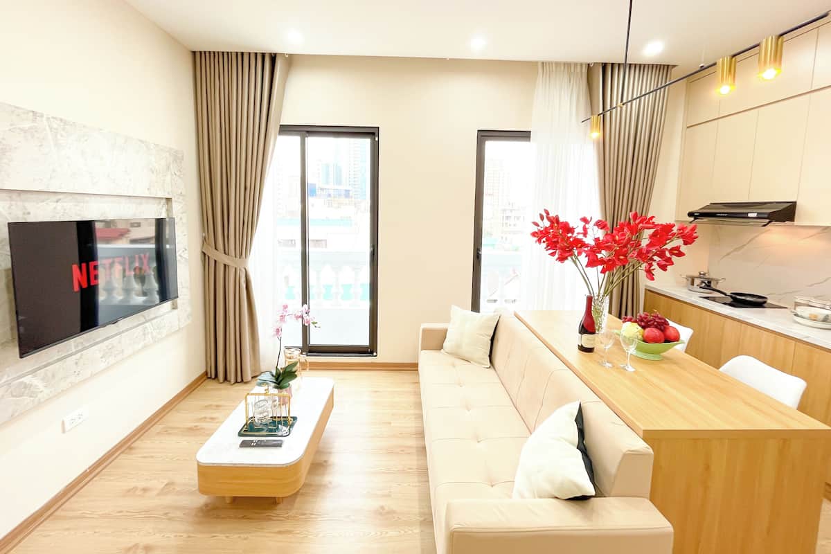 A modern living room with a flat-screen TV, a wooden coffee table, a beige sofa, a dining table with a vase of red flowers, and a kitchen area with a stove and cabinets.
